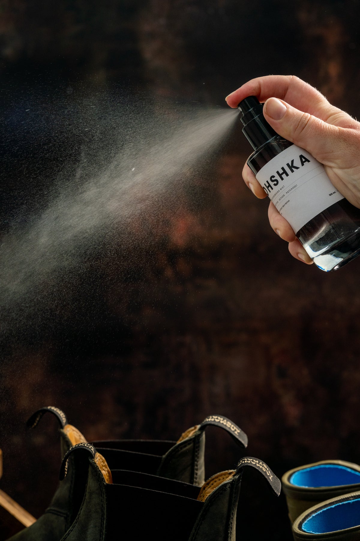 A hand spraying OHSHKA Footwear Refresh Mist into a shoe, the fine mist visible against a dark background